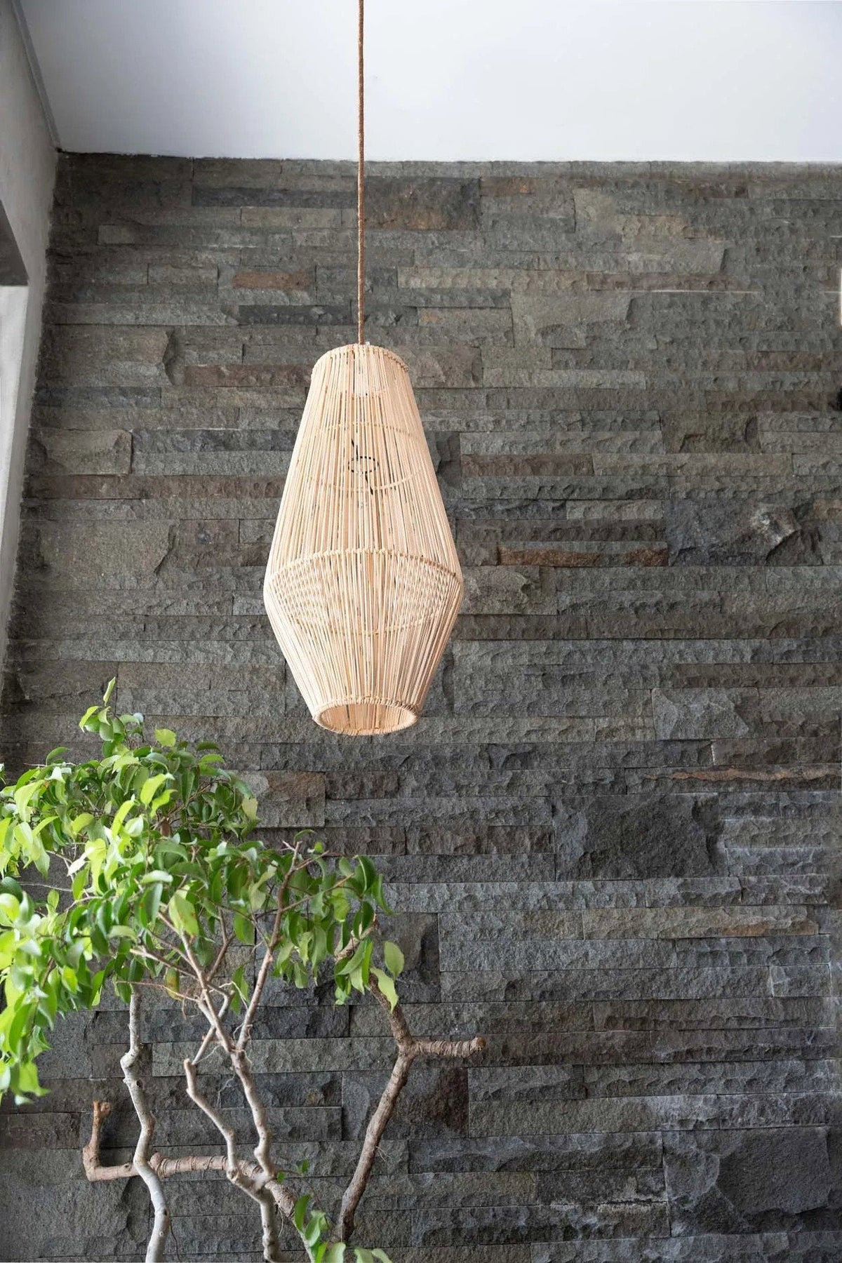 Afrodisiac Rattan Pendant Light – Natural, Medium – suspended against a dark stone wall