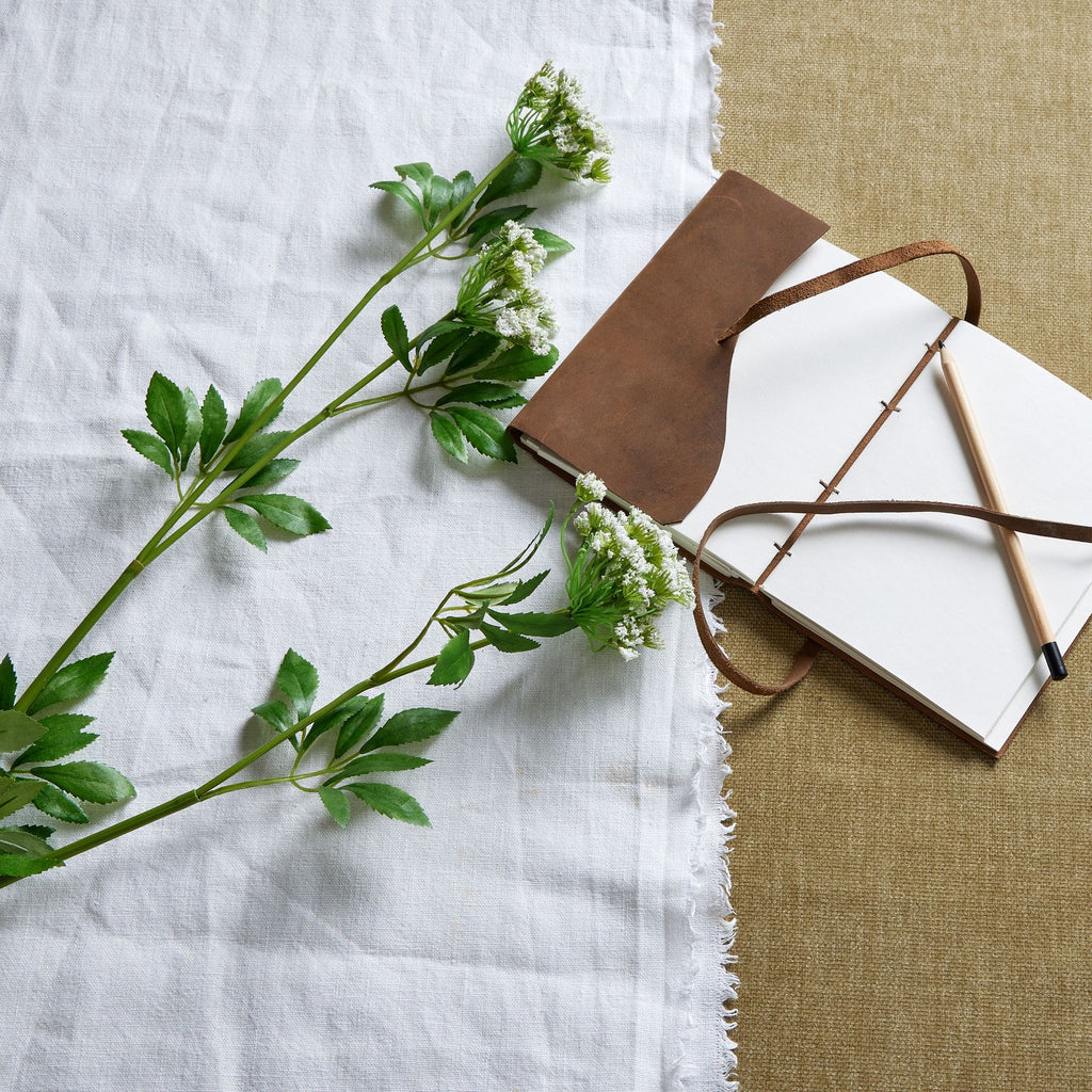 White-flowered artificial sprays laid across a white cloth beside an open brown-covered notebook and pencil.