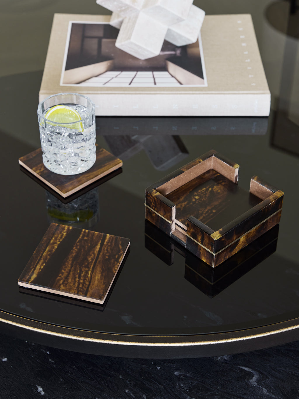 Amber banded coasters and holder on a dark reflective table, with a lemon drink on one coaster and a book behind.
