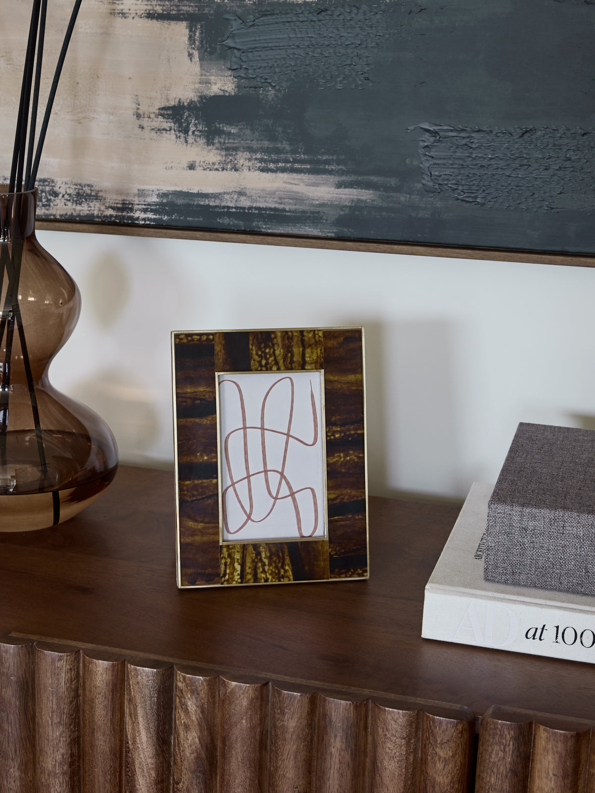 Amber banded photo frame holding a line drawing on a wooden console beside a brown glass vase and stacked books, beneath abstract artwork.