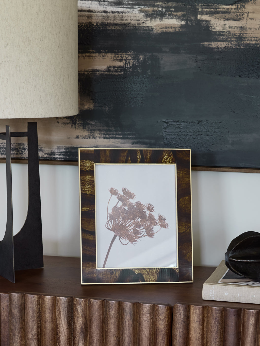 Amber and brown photo frame displaying a botanical print on a wooden console beside a lamp, books and a dark ornament.