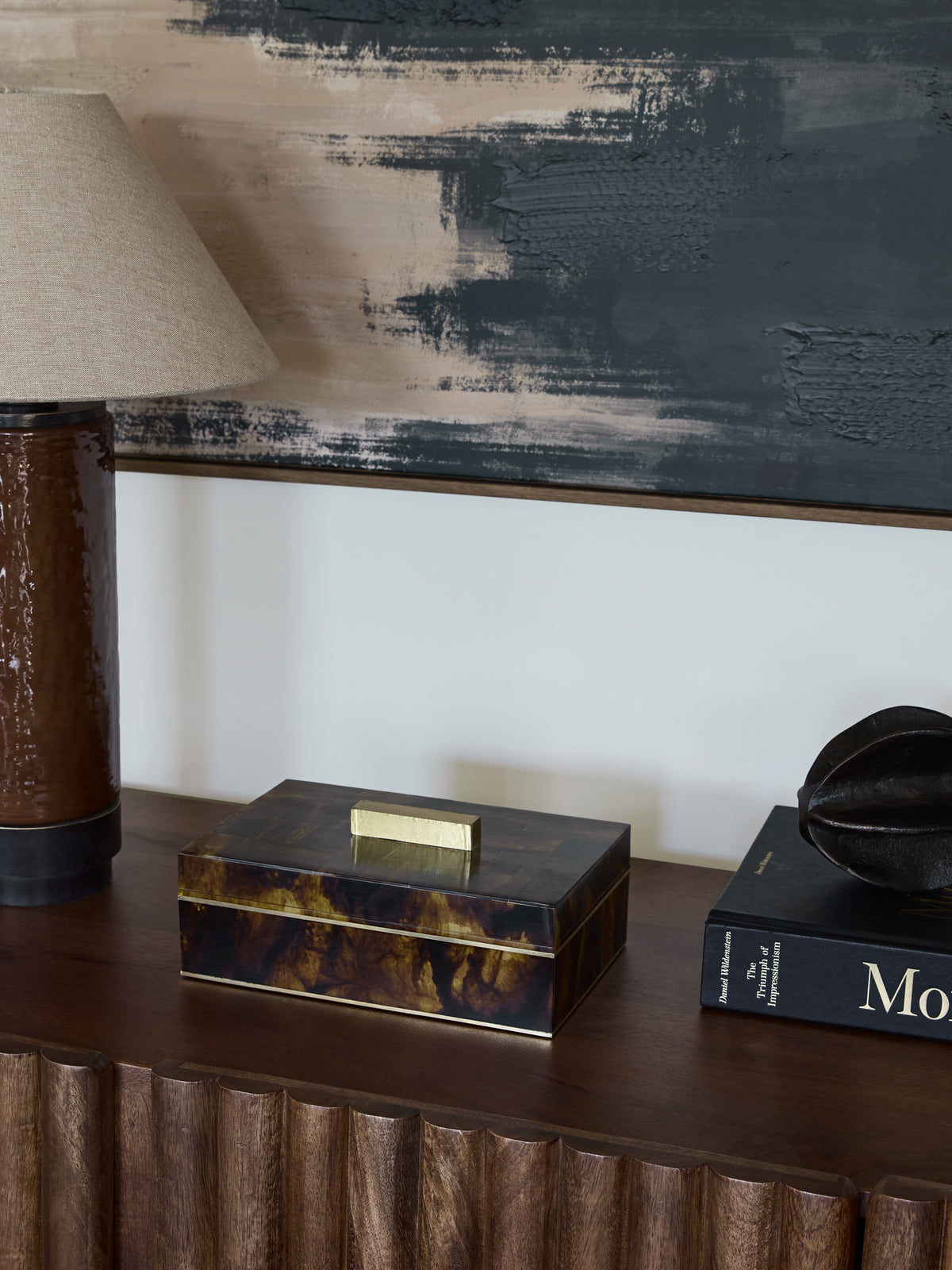 Closed Amber Band Storage Box on a wooden sideboard beside a brown lamp and books, beneath abstract artwork.