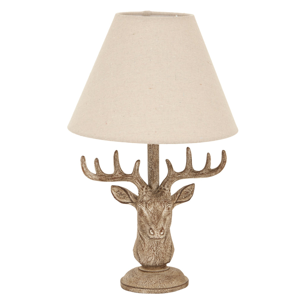 Front view of a stag head table lamp with wide brown antlers, a stepped circular foot and a pale tapered shade on a white background.