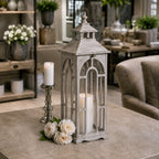 Single pale grey lantern displayed on a table beside a polished candlestick and white flowers, with a lit pillar candle inside.