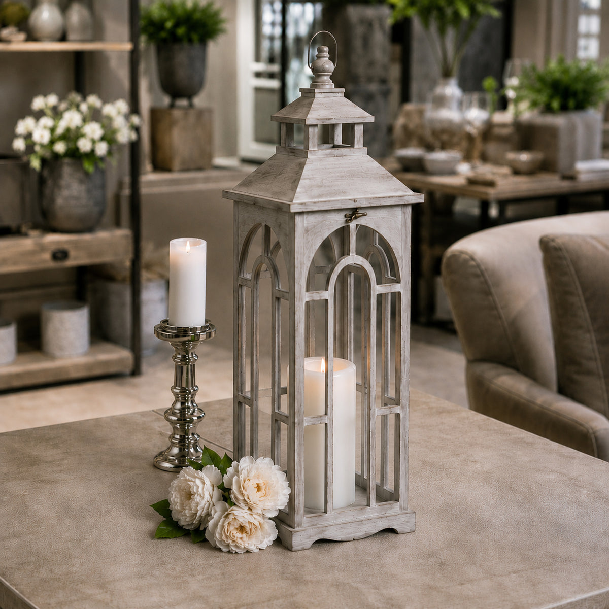 Single pale grey lantern displayed on a table beside a polished candlestick and white flowers, with a lit pillar candle inside.
