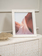 White patterned photo frame holding a pink canyon image on a matching patterned cabinet beside a gold-toned decorative dish.
