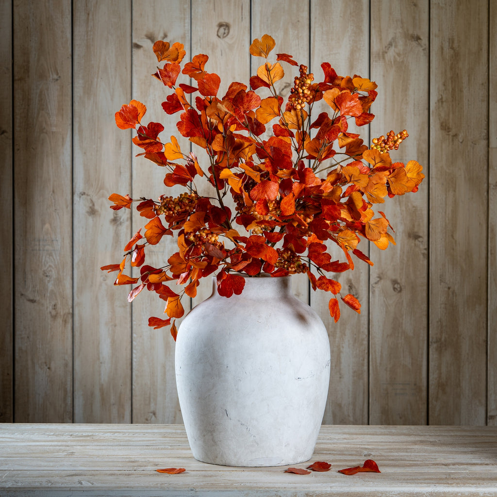 Full autumn foliage arrangement styled in a pale rounded vase on a wooden surface, with loose leaves below and timber panelling behind.