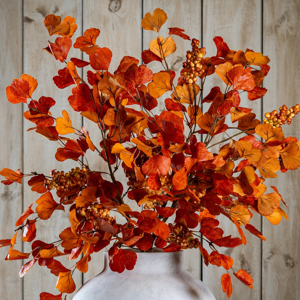 Closer view of orange and burgundy fan-shaped foliage and berry clusters spreading above the neck of a pale vase against wooden panelling.