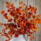 Closer view of orange and burgundy fan-shaped foliage and berry clusters spreading above the neck of a pale vase against wooden panelling.