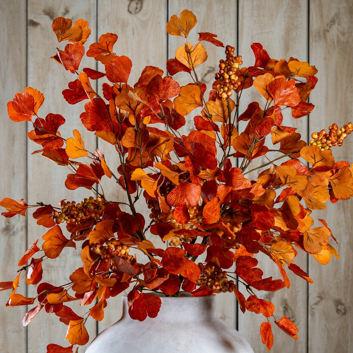 Closer view of orange and burgundy fan-shaped foliage and berry clusters spreading above the neck of a pale vase against wooden panelling.