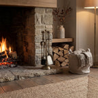 Yellow-brown fireside companion set on a stone hearth beside a lit fireplace, with stacked logs, a vase of branches and a basket holding a knitted throw nearby.
