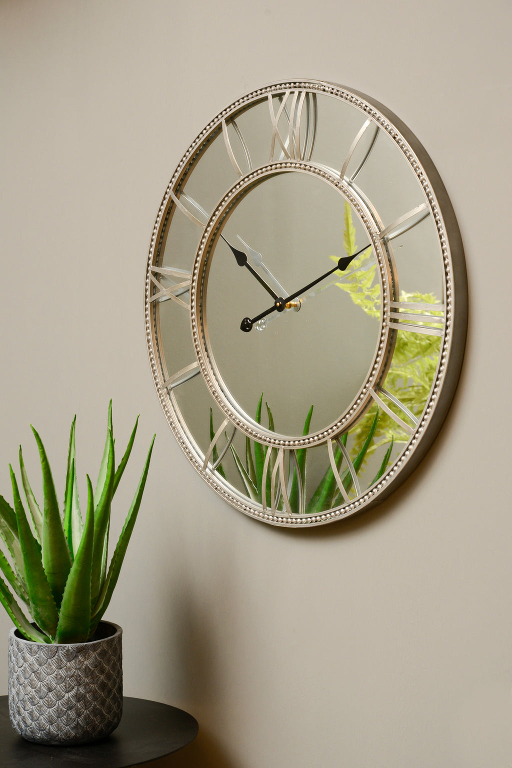 Angled view of the beaded mirrored wall clock on a neutral wall above a black table, with a potted aloe reflected in the dial.