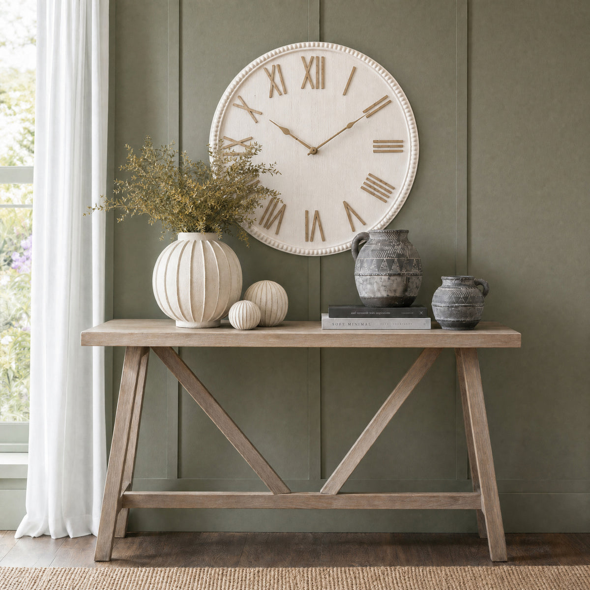 Beaded Roman wall clock on a muted green panelled wall above a wooden console styled with pale ornaments, foliage and dark patterned vessels.
