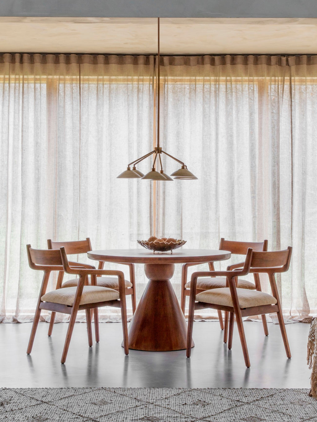 Bell shade chandelier suspended above a round wooden dining table and four upholstered wooden chairs in front of sheer curtains.