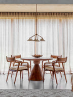 Bell shade chandelier suspended above a round wooden dining table and four upholstered wooden chairs in front of sheer curtains.