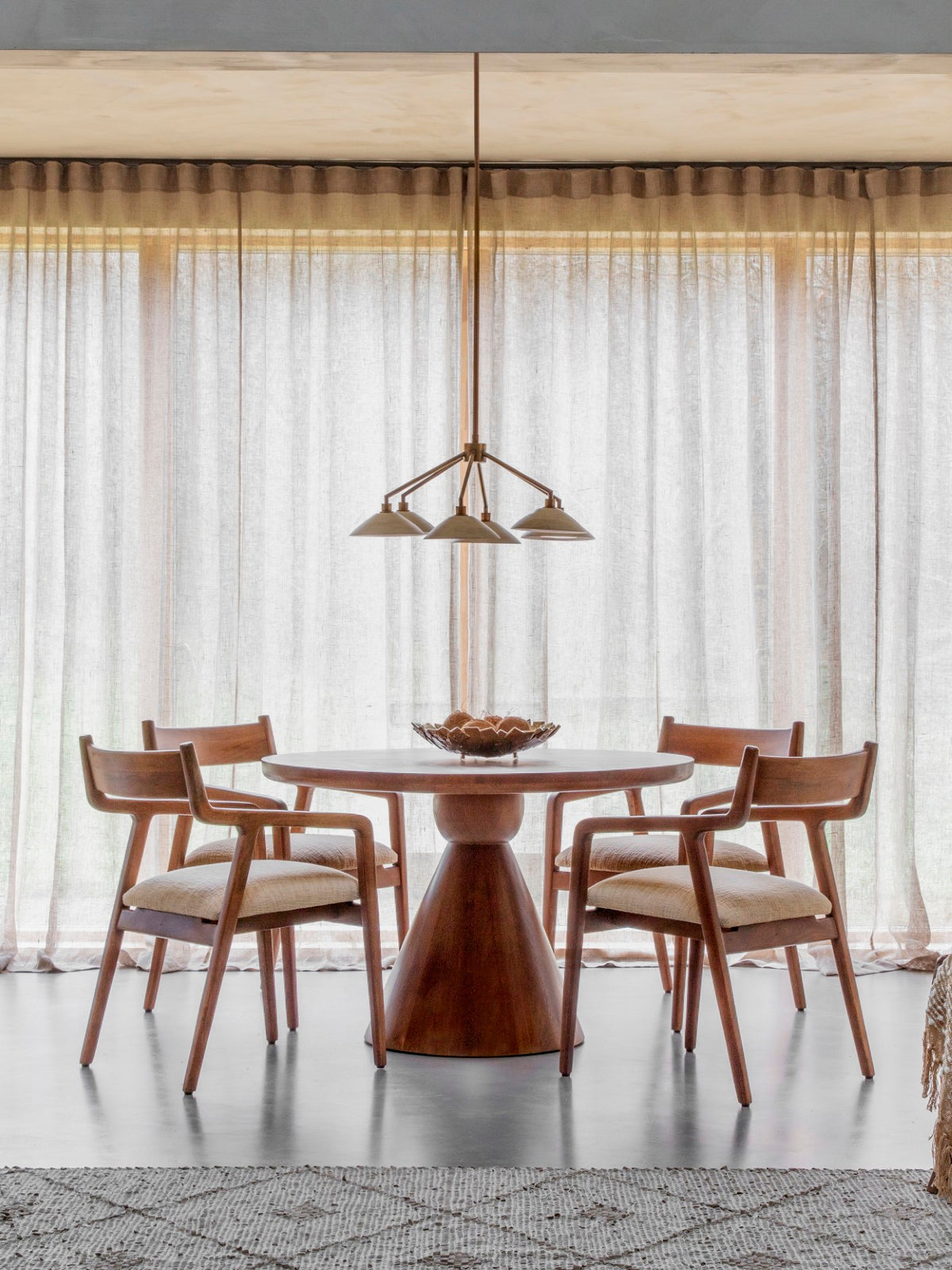 Bell shade chandelier suspended above a round wooden dining table and four upholstered wooden chairs in front of sheer curtains.