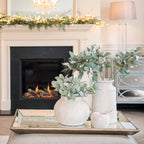 Mirrored rectangular tray styled with two pale vases of leafy stems and a decorative apple on an upholstered surface in front of a fireplace.