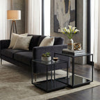 Two black framed side tables beside a charcoal sofa, styled with books, a flower vase, a gold ornament, a small plant and a cup on a pale rug.