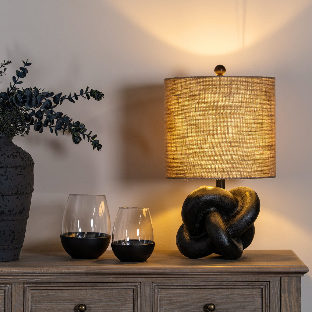Illuminated Black Interlock Table Lamp on a wooden cabinet beside two glass vessels and a tall vase of leafy stems.