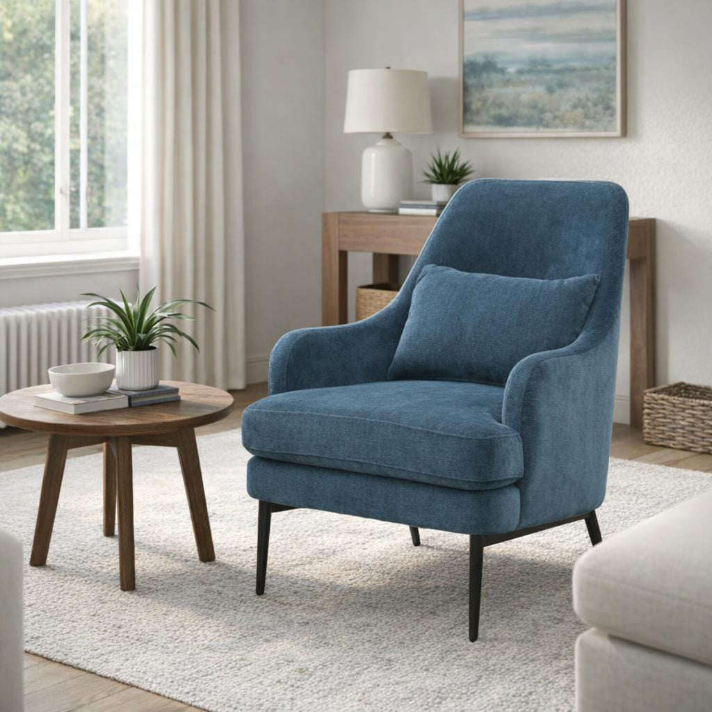 Blue armchair on a pale rug beside a round wood table, with a console, lamp and framed artwork behind it.