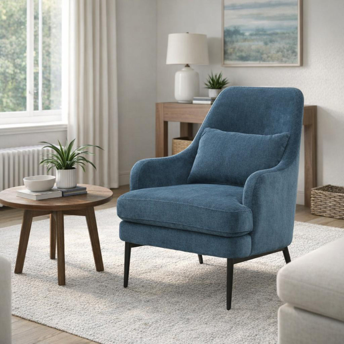Blue armchair on a pale rug beside a round wood table, with a console, lamp and framed artwork behind it.