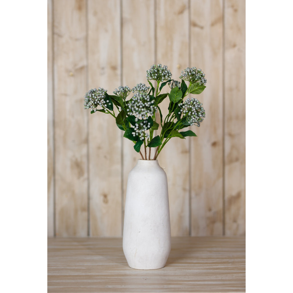 Blue-grey hydrangea bud sprays displayed in a tall white vase on a wooden surface against vertical wood panelling.