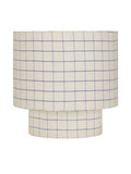 Front view of a table lamp with a broad drum shade and short cylindrical base, both covered in natural-toned fabric with fine blue checks, on white.