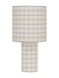 Table lamp on a white background with a broad cylindrical shade and slim cylindrical base, both covered in natural-toned fabric with a fine blue check.