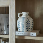 Blue leaf vase with its handle facing left, displayed on a large pale book inside a wooden bookcase beside upright books.