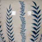 Close-up of alternating dark and pale blue leafy stems over glossy off-white glaze, showing the fine crackle pattern and scattered beige-brown marks.