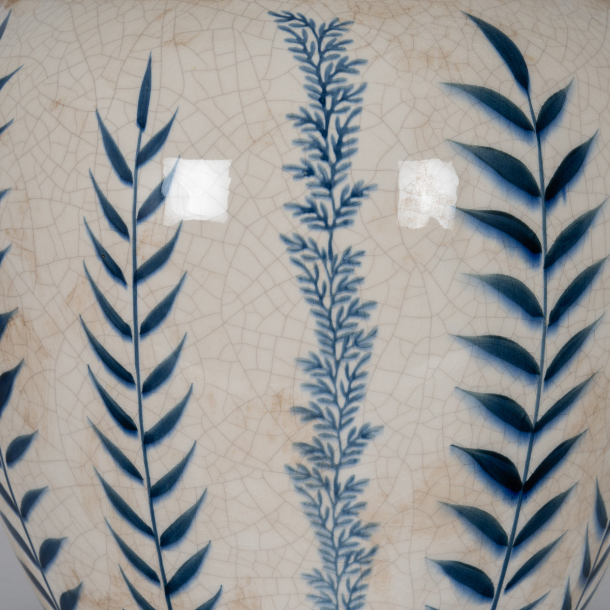 Close-up of alternating dark and pale blue leafy stems over glossy off-white glaze, showing the fine crackle pattern and scattered beige-brown marks.