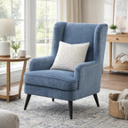 Blue wing armchair styled with a cream cushion beside a wooden side table and lamp, on a patterned rug by large windows.