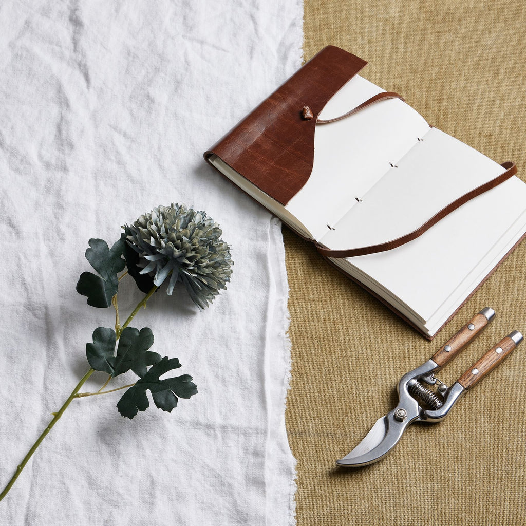 Artificial blue-green flower stem laid diagonally on white cloth beside an open brown notebook and wooden-handled secateurs on a tan surface.