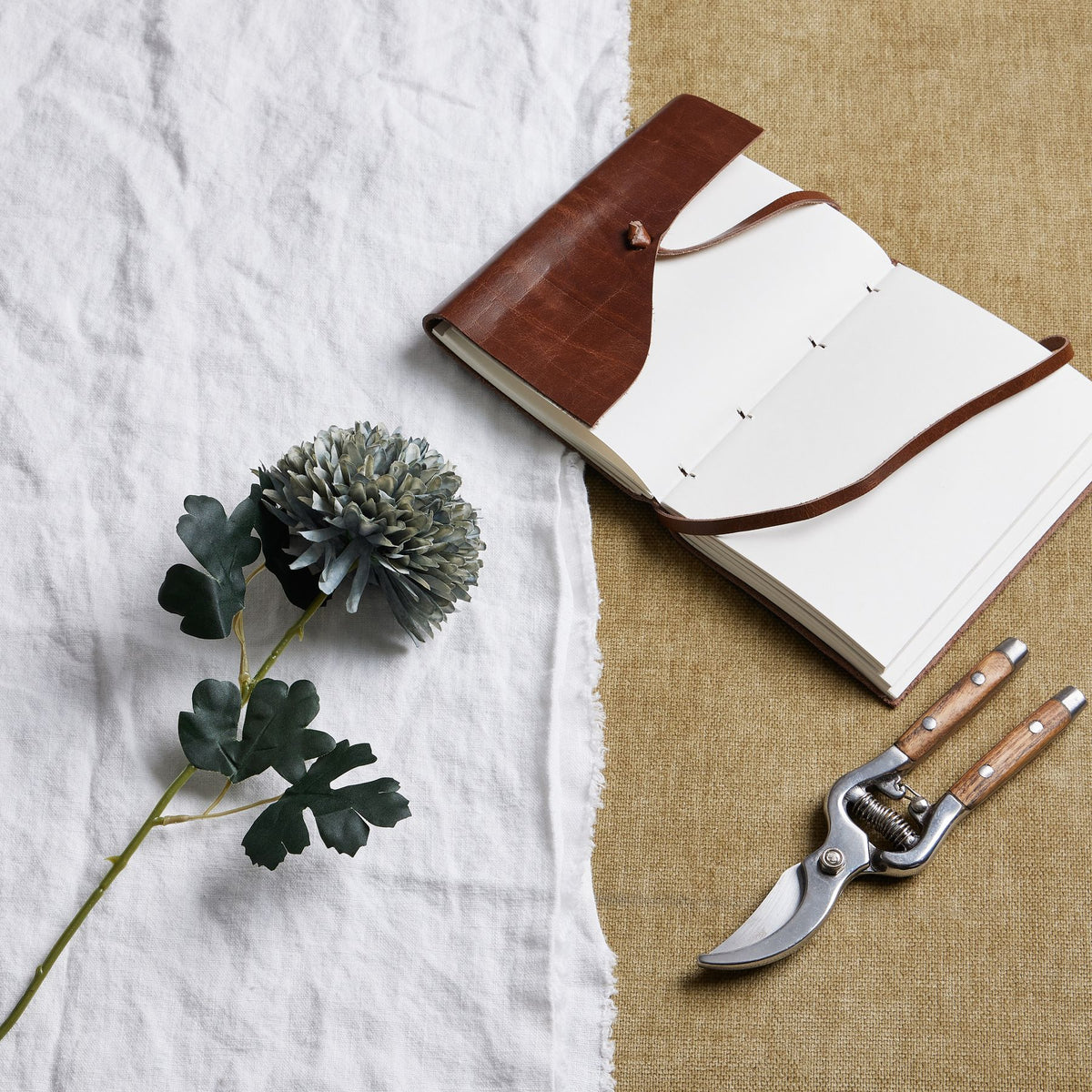 Artificial blue-green flower stem laid diagonally on white cloth beside an open brown notebook and wooden-handled secateurs on a tan surface.