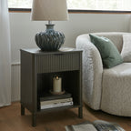 Bluegreen Waveflute Table Lamp on a dark side table beside a cream armchair with a green cushion, with books and a candle on the lower shelf.