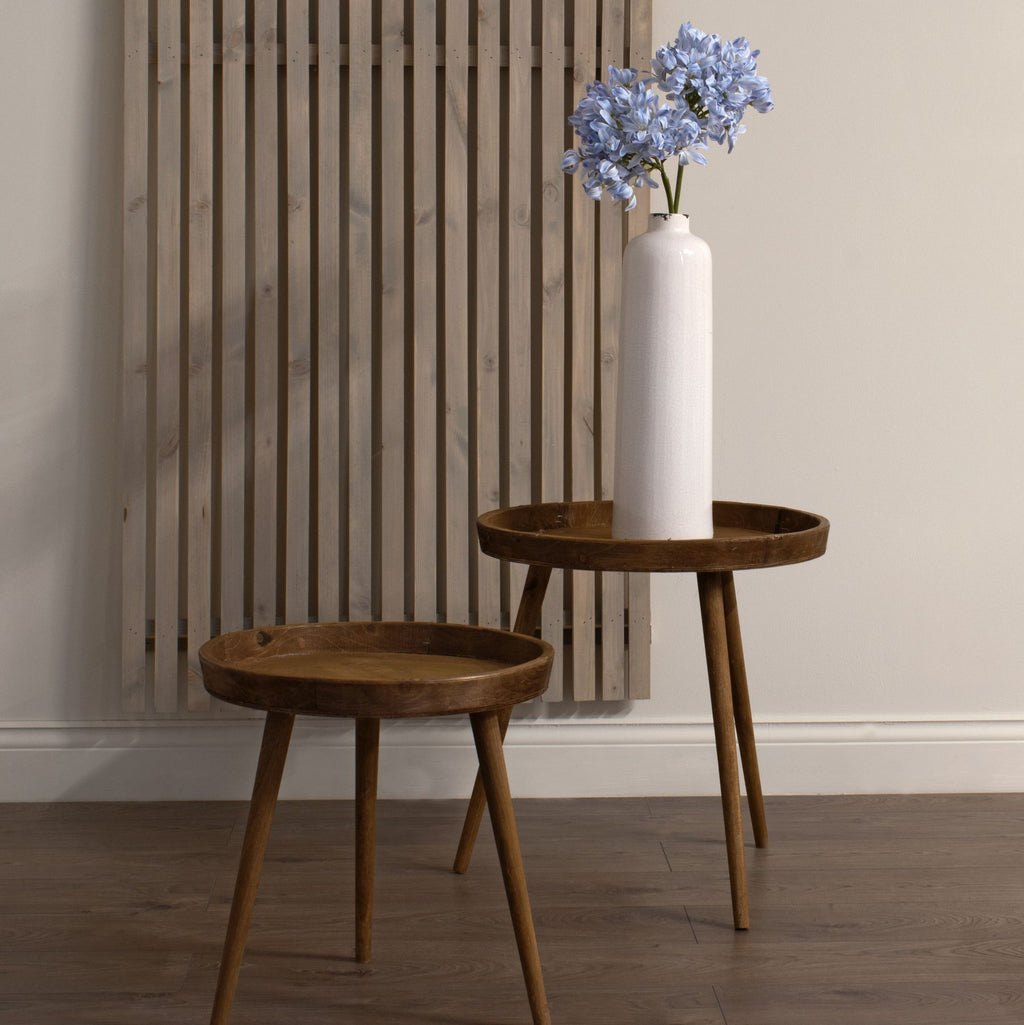 Pale blue artificial flower stems arranged in a tall white vase on a wooden tray table, beside a lower empty table and a vertical slatted wall panel.