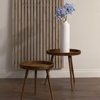 Pale blue artificial flower stems arranged in a tall white vase on a wooden tray table, beside a lower empty table and a vertical slatted wall panel.