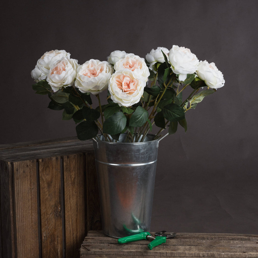 Several ivory and blush artificial flowers arranged in a tall metal bucket on rustic wooden crates, with green-handled secateurs in front.