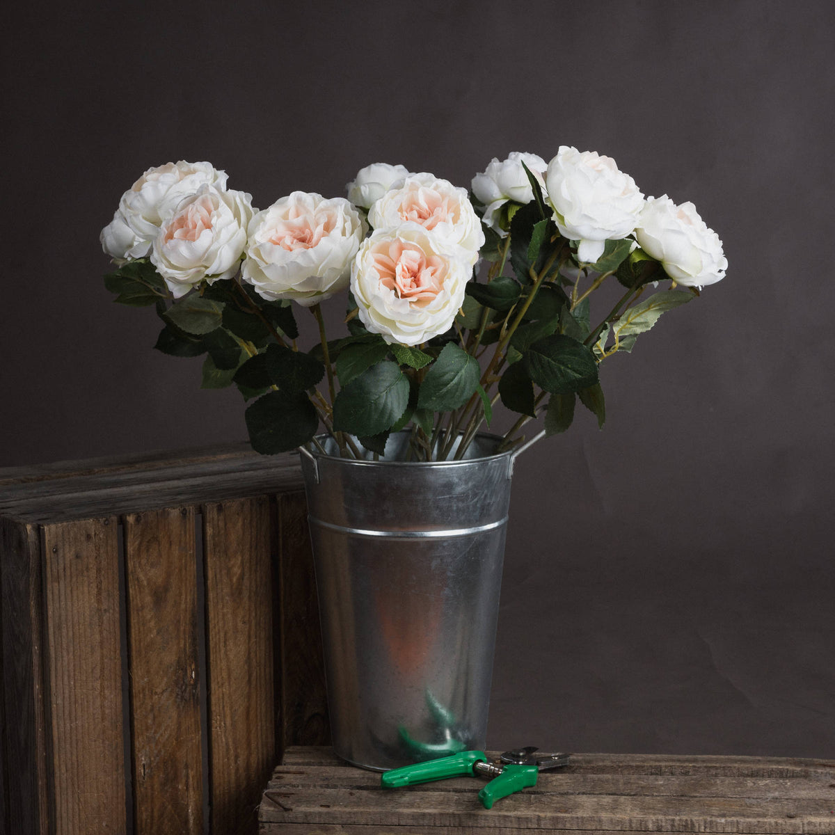 Several ivory and blush artificial flowers arranged in a tall metal bucket on rustic wooden crates, with green-handled secateurs in front.