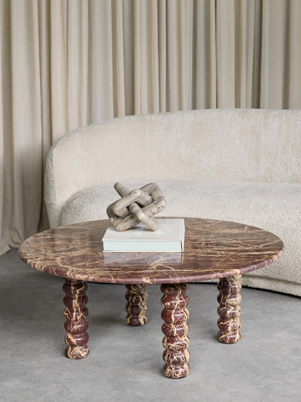 Bobbin-leg marble coffee table styled with a book and interlocking sculpture in front of a cream curved sofa and curtains.