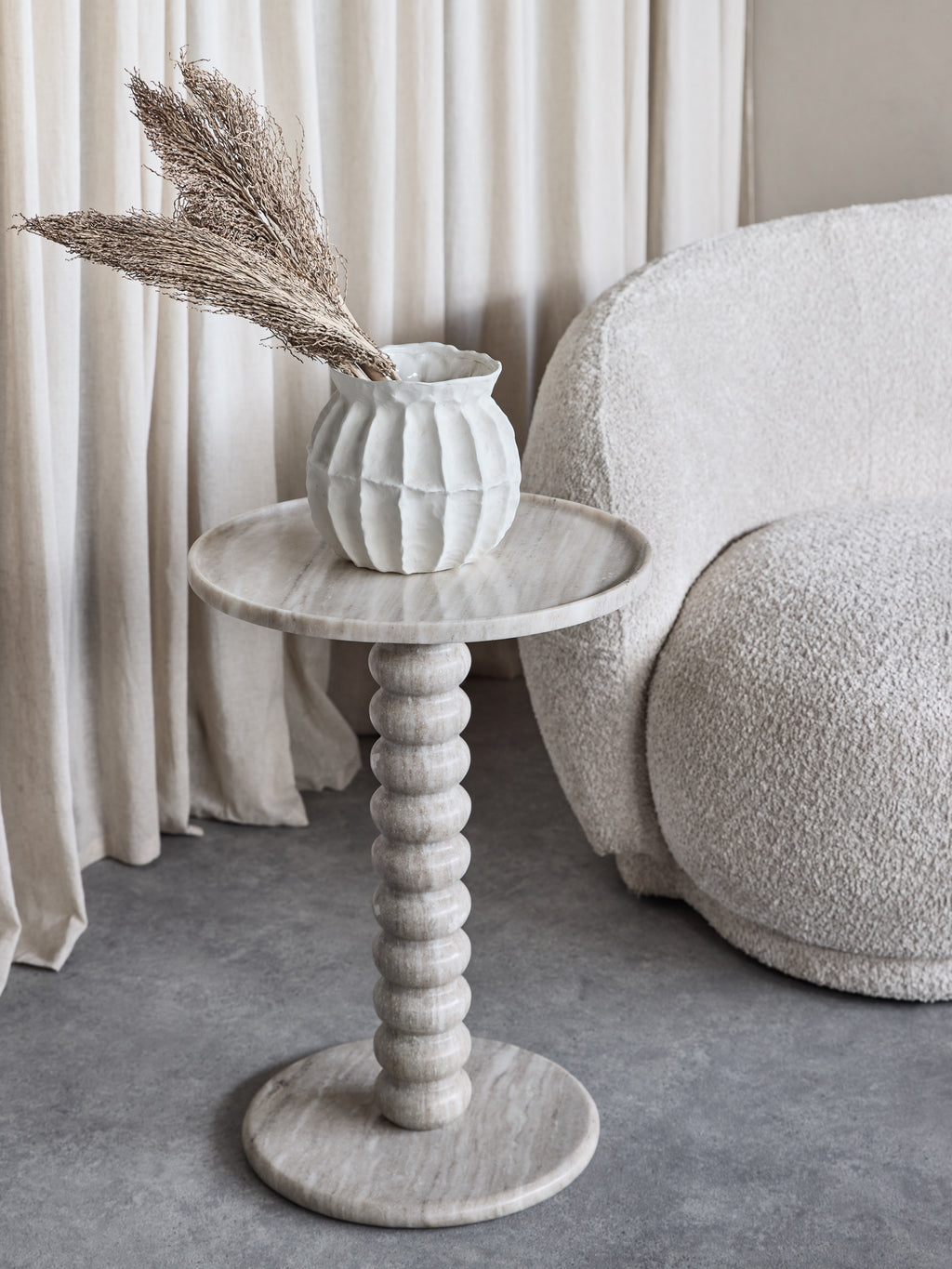 Beige marble bobbin side table beside a cream textured armchair, styled with a white vase and dried stems in front of pale curtains.