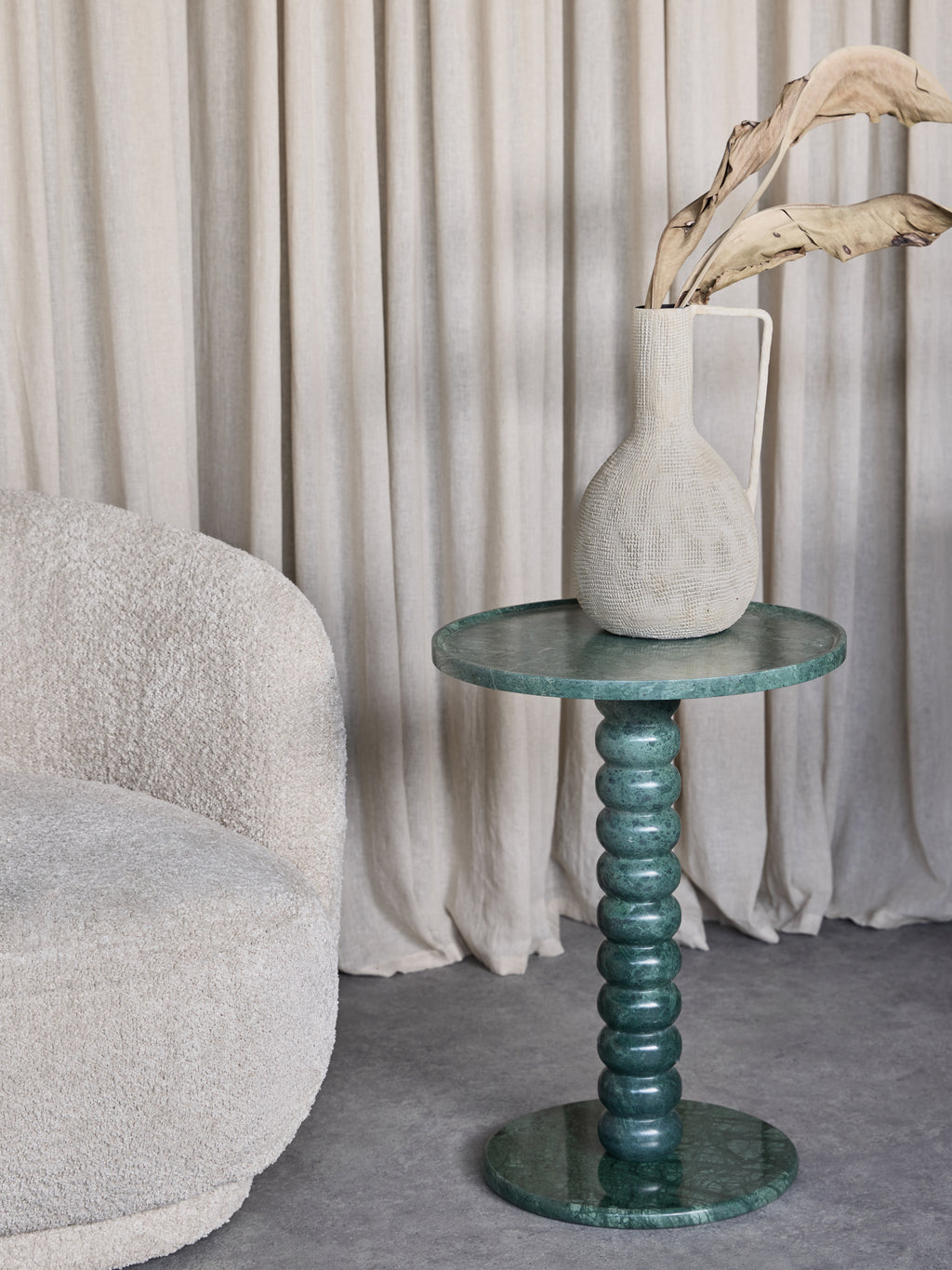 Green marble bobbin side table beside a cream textured armchair, topped with a pale jug holding dried leaves, with curtains behind.