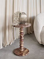 Red marble bobbin side table beside a cream textured chair and pale curtains, styled with a metallic vase and trailing stems.