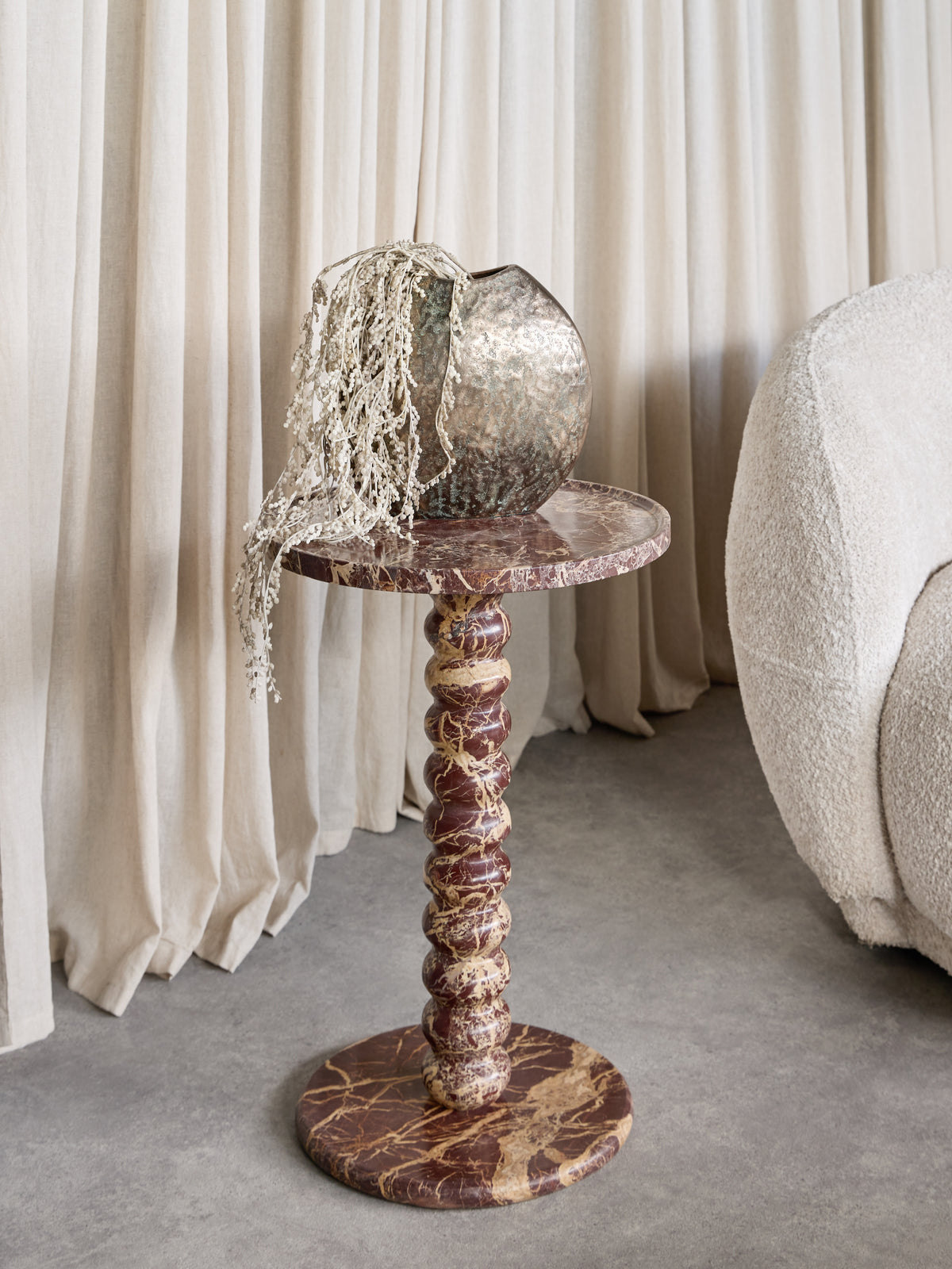 Red marble bobbin side table beside a cream textured chair and pale curtains, styled with a metallic vase and trailing stems.