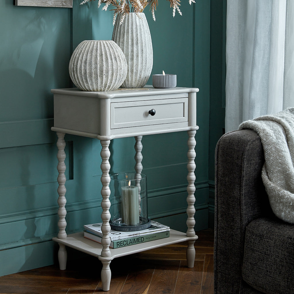 Pale grey bobbin side table beside a grey armchair against teal panelling, styled with ribbed vases above and books on the lower shelf.