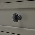 Dark round drawer knob against the pale grey drawer front, with fine surface grain and raised framing visible.