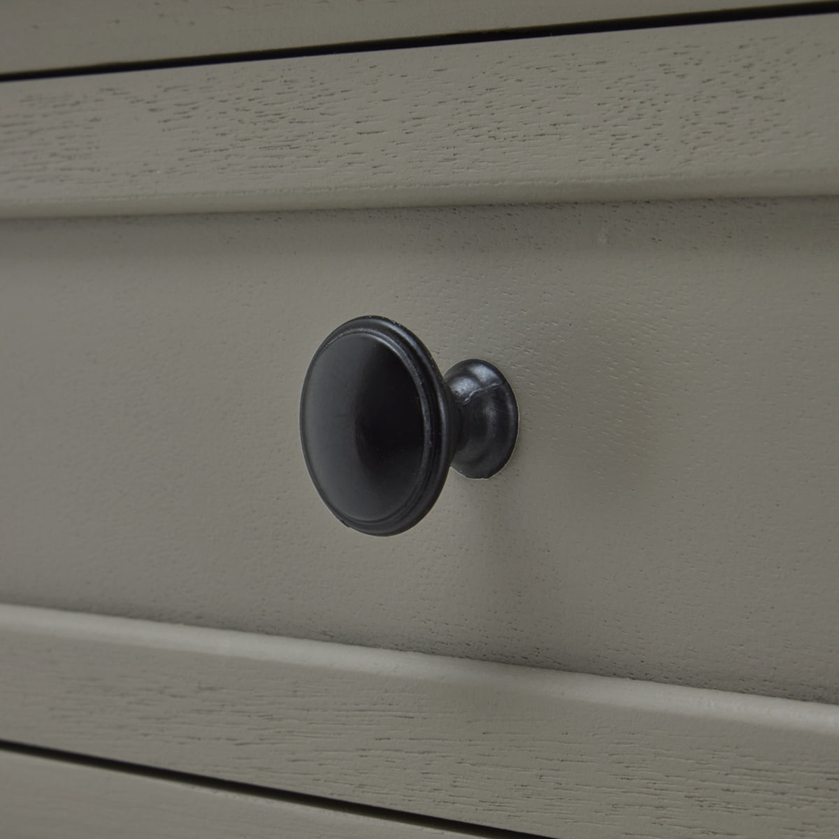 Dark round drawer knob against the pale grey drawer front, with fine surface grain and raised framing visible.