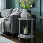Two-tier bobbin side table beside a grey sofa and teal panelled wall, styled with a vase of green stems, candles and stacked magazines.