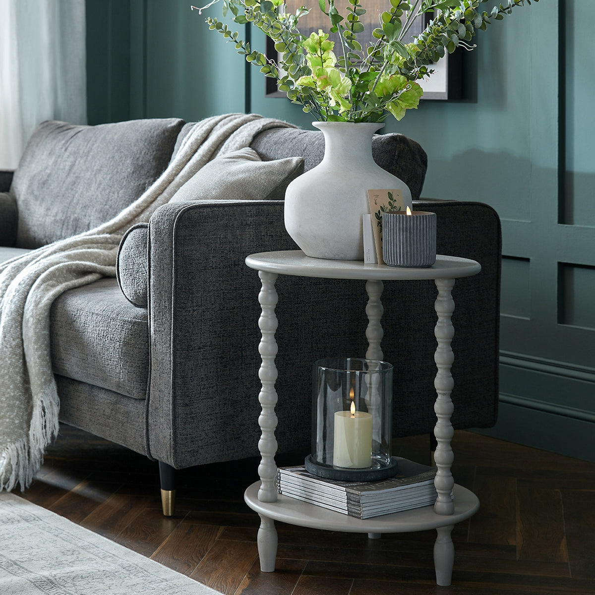 Two-tier bobbin side table beside a grey sofa and teal panelled wall, styled with a vase of green stems, candles and stacked magazines.