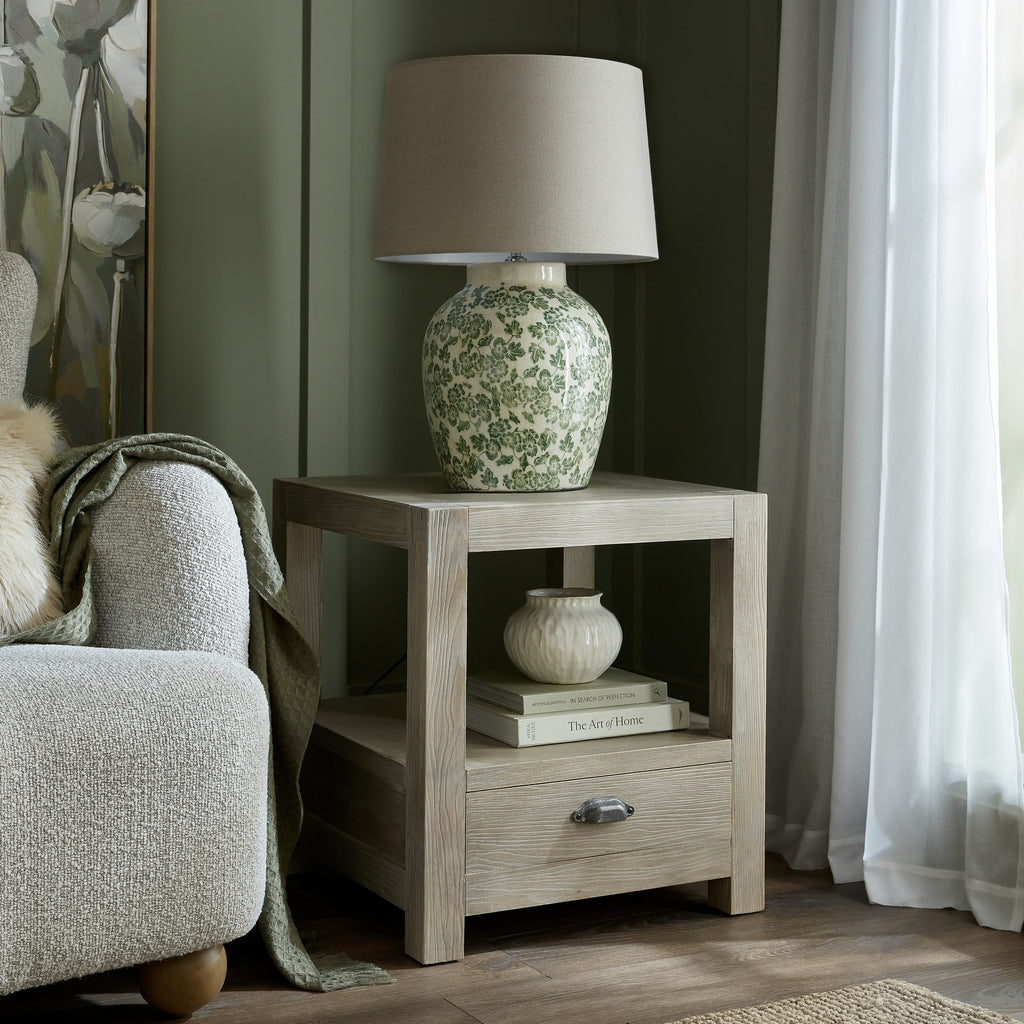 Side table beside a cream armchair, styled with a green floral lamp on top and a small cream vase on books on the open shelf.